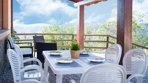Un patio al aire libre con una mesa dispuesta para dos personas, rodeado de sillas blancas, con vistas a un paisaje montañoso.