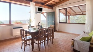 Un comedor con mesa y sillas de madera, un televisor montado en la pared y ventanas amplias con vistas al exterior.