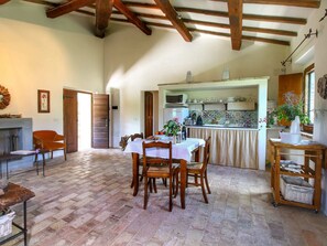A rustic dining area with a wooden table and chairs, a fireplace, and a kitchen in the background.