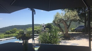 Ett bord med ett glas vitt vin och en vas, placerat under ett parasoll, med utsikt över ett vackert landskap med träd och en bil.