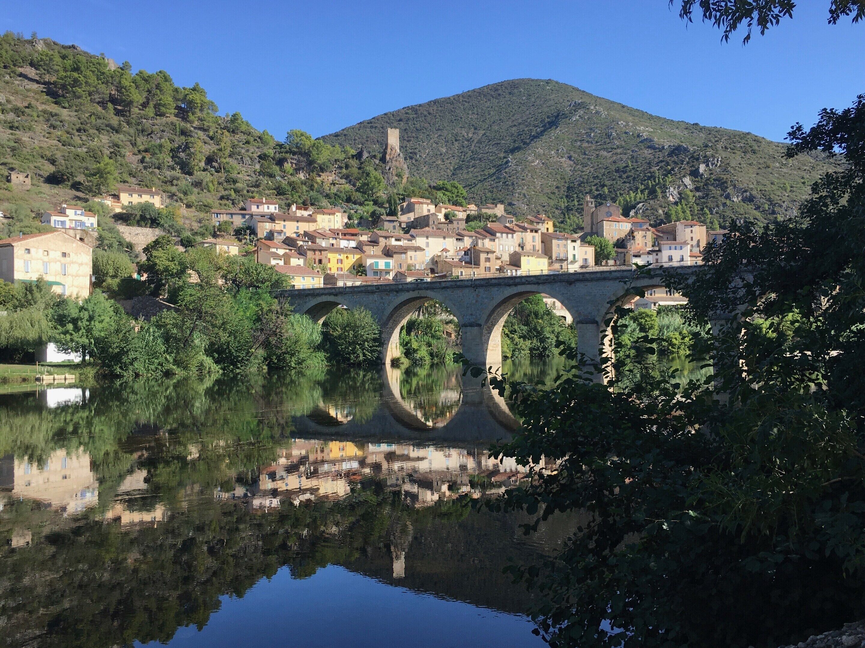 Un pont à trois arches enjambant une rivière paisible, entouré d’une végétation luxuriante et surplombant un village perché sur une colline, avec ses bâtiments.
