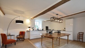 A spacious room with a wooden dining table, a sofa, and a chair. There is a kitchen area in the background with a window. - Holiday Home in Angers With Private Garden (Bierné-les-Villages)