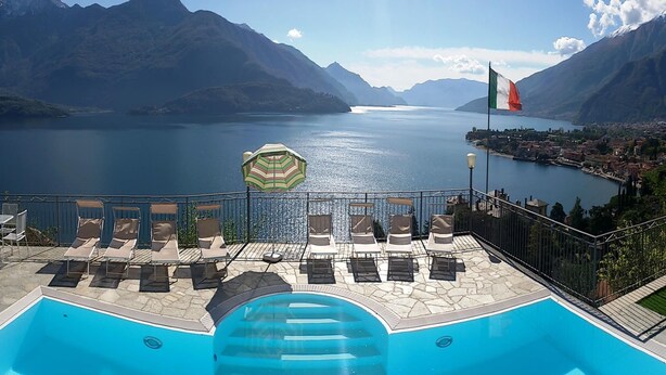 A swimming pool with lounge chairs and an umbrella, overlooking a lake with mountains in the background.