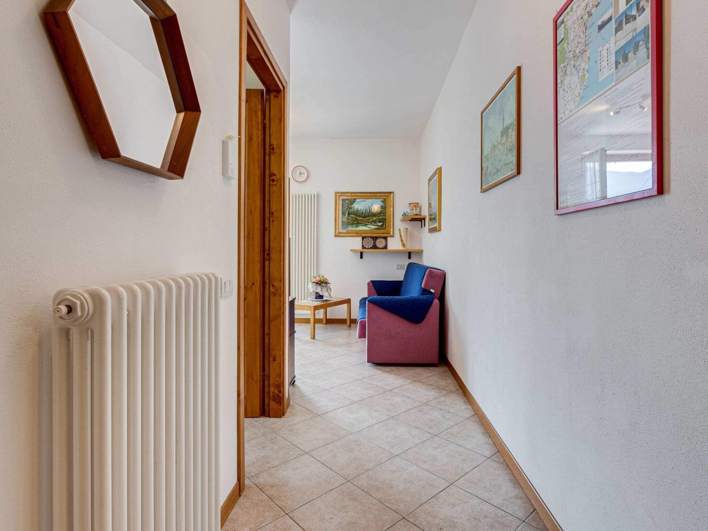 A hallway with a radiator, a wooden door, a blue and red chair, a small table with a vase, and a framed picture on the wall.