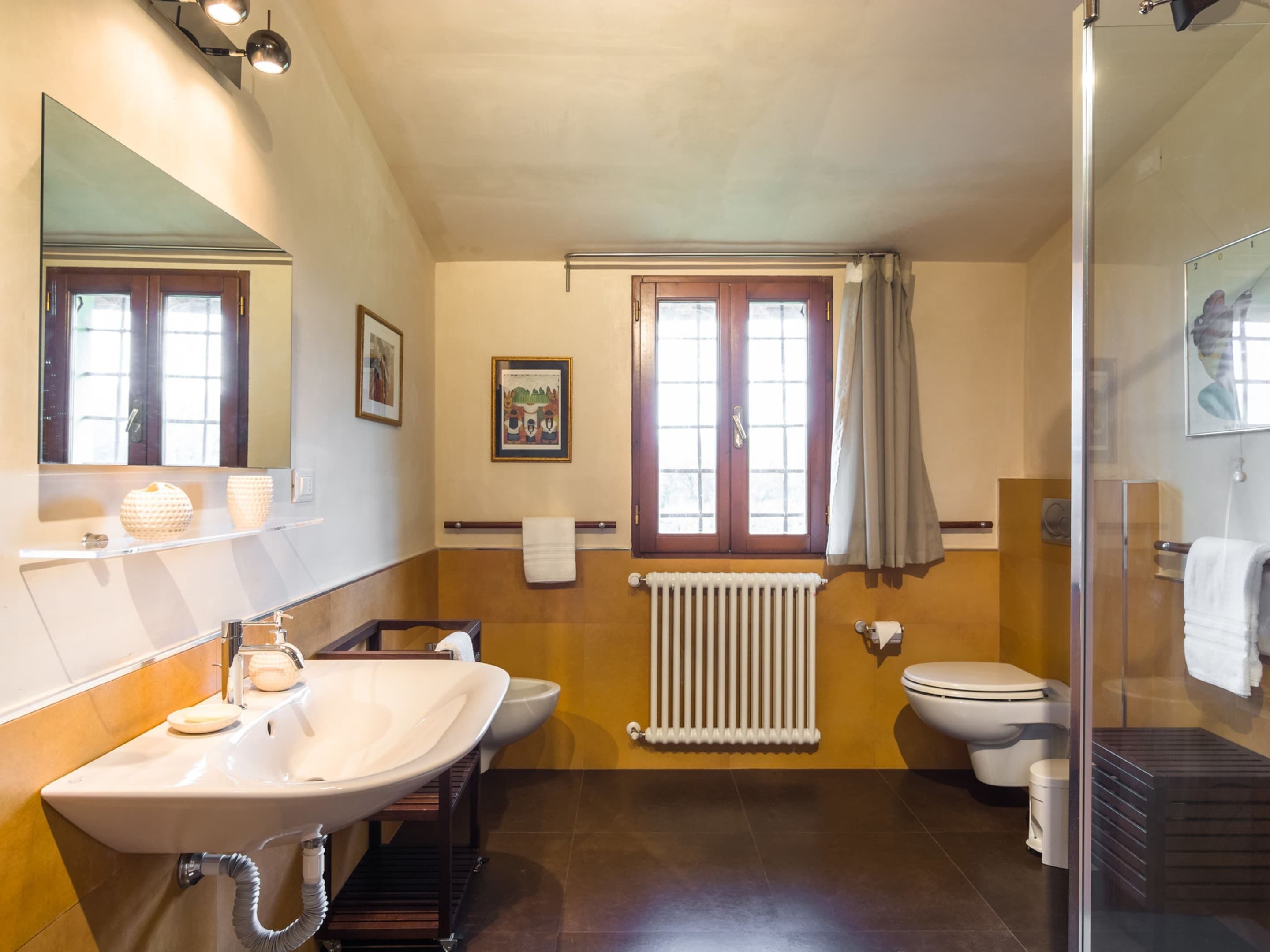 A bathroom with a sink, toilet, and shower. There is a window with curtains, a radiator, and a towel rack.