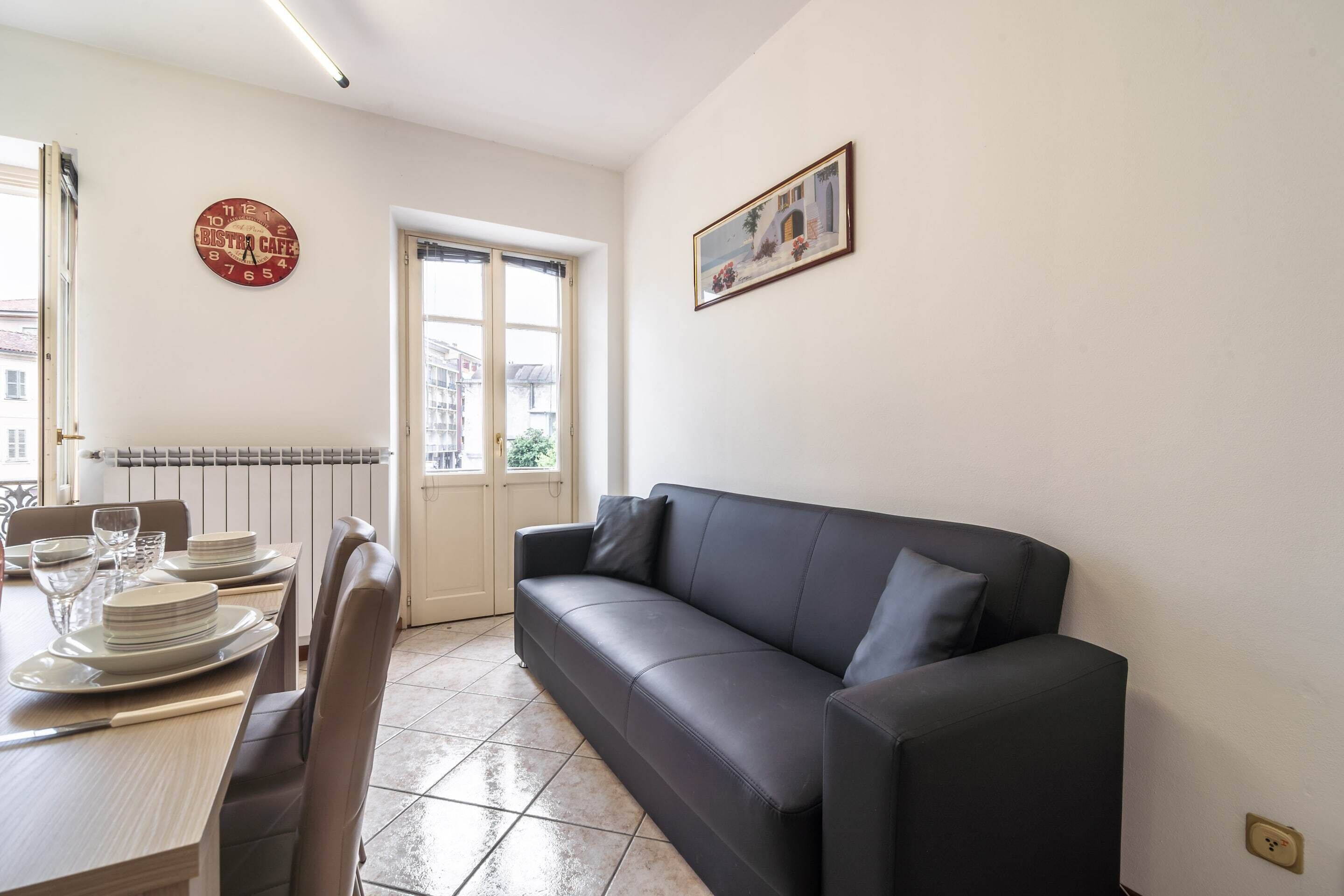 Een eetruimte met een tafel gedekt voor vier personen, een zwarte leren bank, een klok en een lijst met een afbeelding aan de muur.