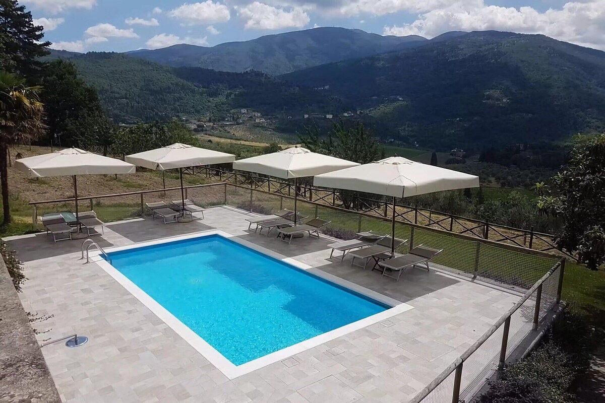A rectangular pool with a clear blue water surface, surrounded by lounge chairs and umbrellas, with a view of green mountains in the background.