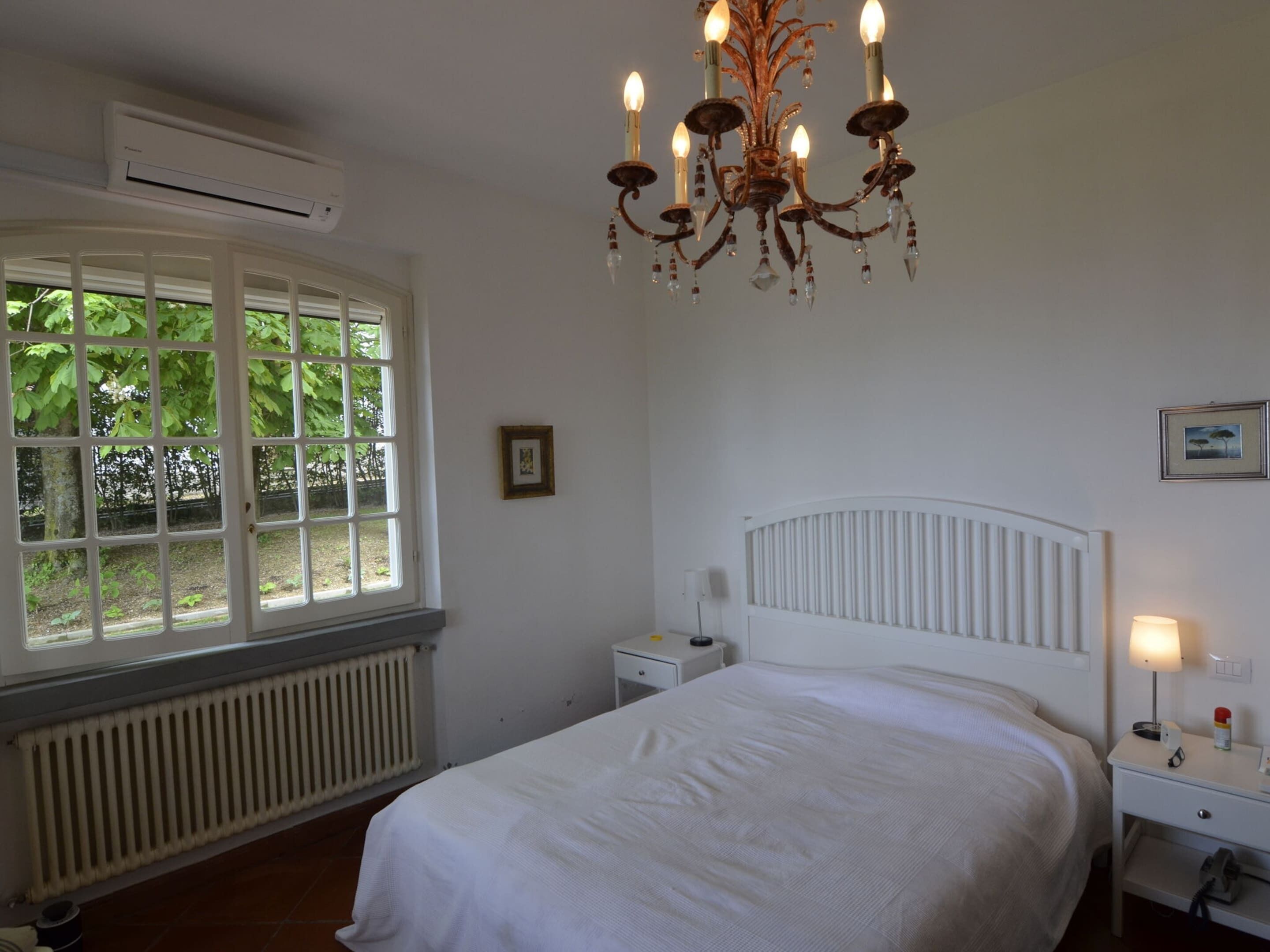 Quarto com cama branca, mesa de cabeceira branca, radiador branco, janela com vista para área verde e um lustre.