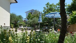 A garden with a white building, a blue and white striped umbrella, and outdoor furniture. - Apartment in Gatteo Mare Near Beach (Gatteo)