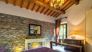 Une chambre avec un mur en pierre, un lit recouvert d’une courtepointe violette, un banc en bois, une commode et un lustre.
