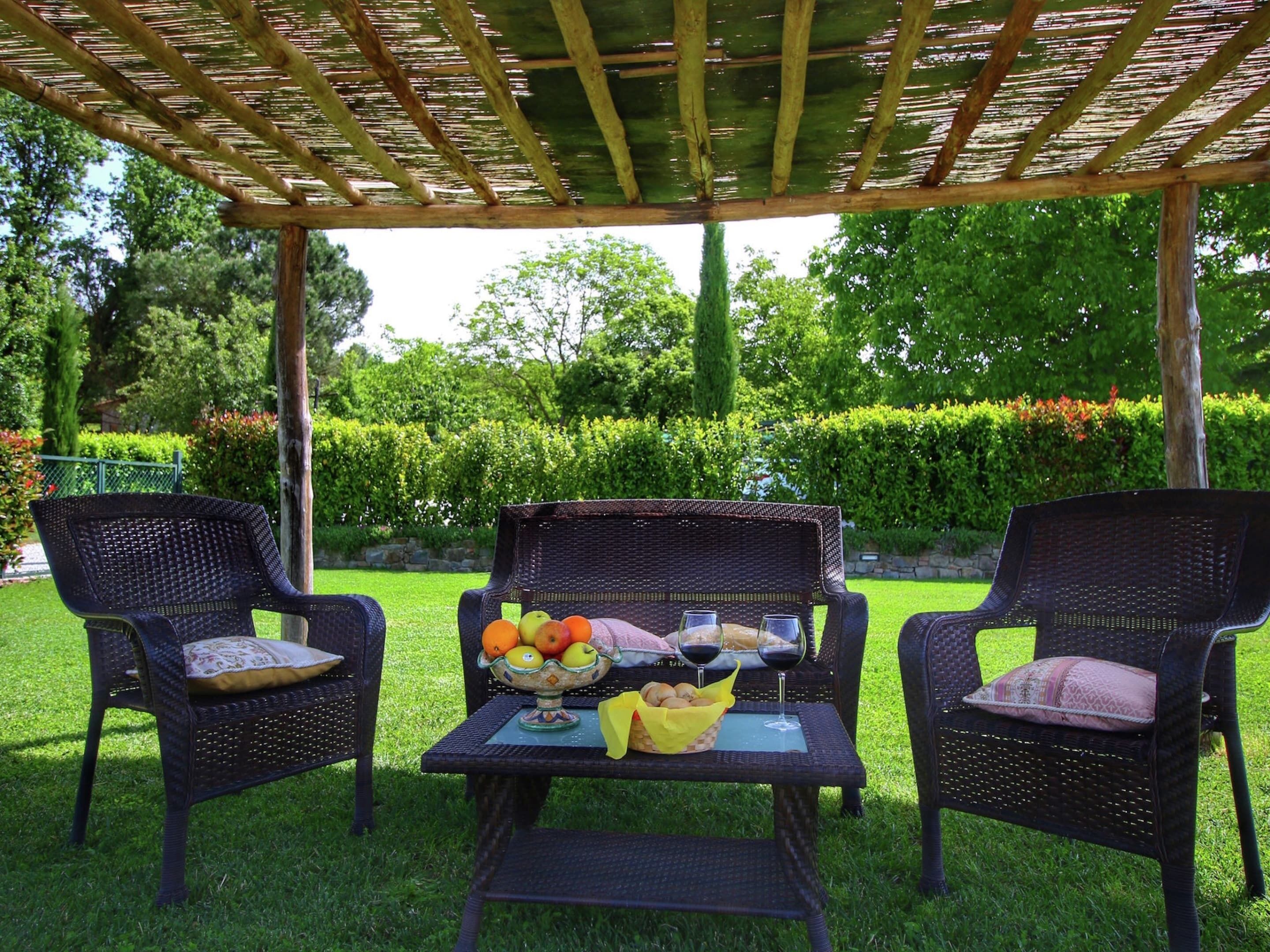 Un área de estar al aire libre con sillas de mimbre y una mesa dispuesta con frutas, pan y copas de vino.
