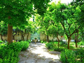 A stone pathway surrounded by lush greenery and trees, leading to a seating area with chairs and a table. - Belvilla by OYO Palazzo Pio (Lecce)