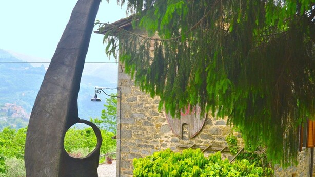 Un bâtiment en pierre doté d’une fenêtre circulaire et d’une porte en bois, entouré d’une végétation luxuriante et offrant une vue sur des montagnes lointaines.