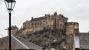 Un bâtiment historique en pierre, doté d’un escalier menant à un niveau inférieur, d’un lampadaire et offrant une vue sur un château perché sur une colline au loin.