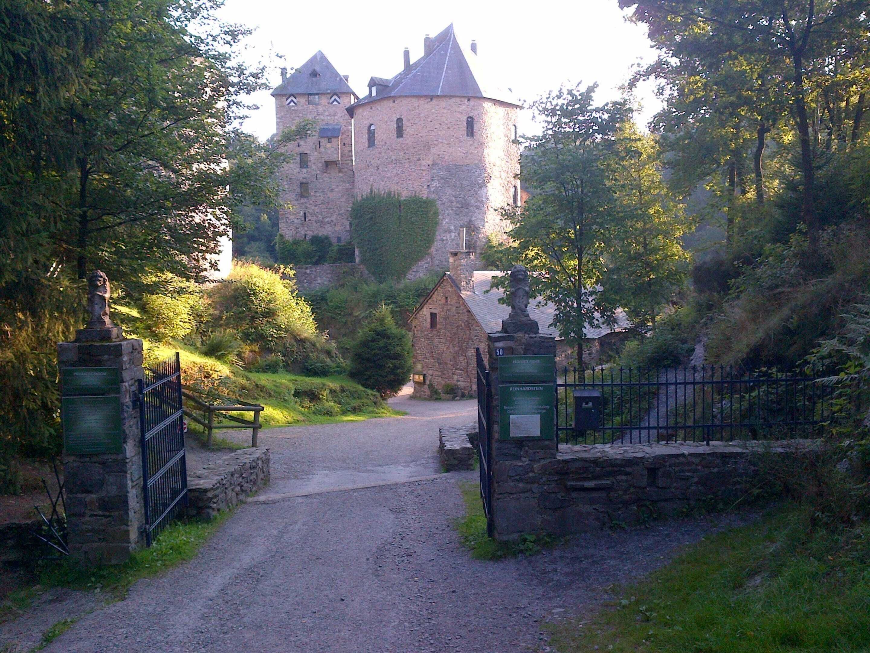 A castle entrance with a gravel path leading to a stone gate, surrounded by trees and greenery.
