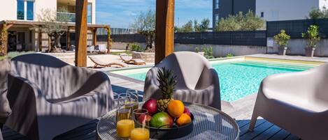 A poolside area with a table set with fruit and drinks, lounge chairs, and a view of a modern building and pool.
