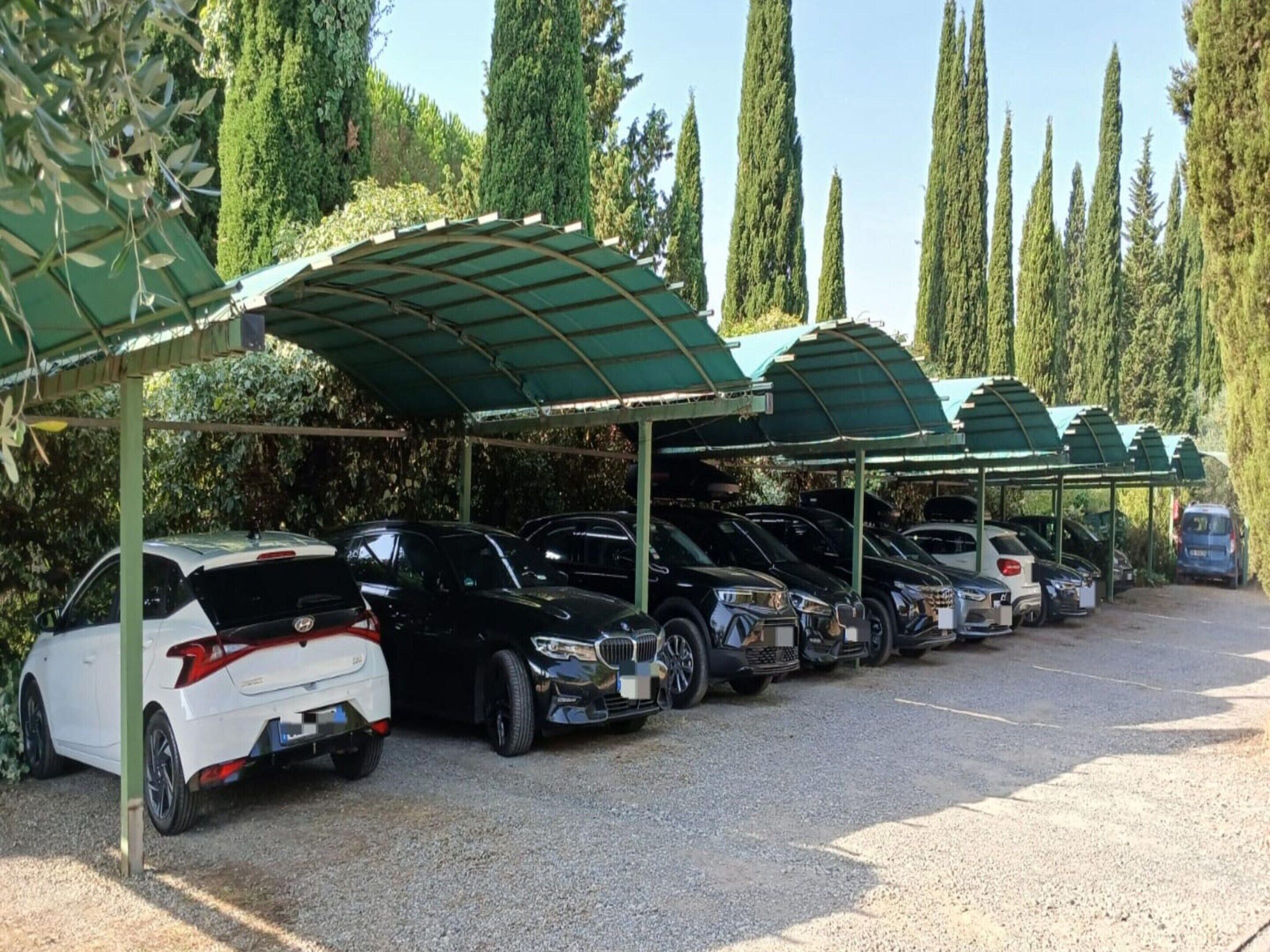A parking area with multiple cars under green canopies, surrounded by tall trees.