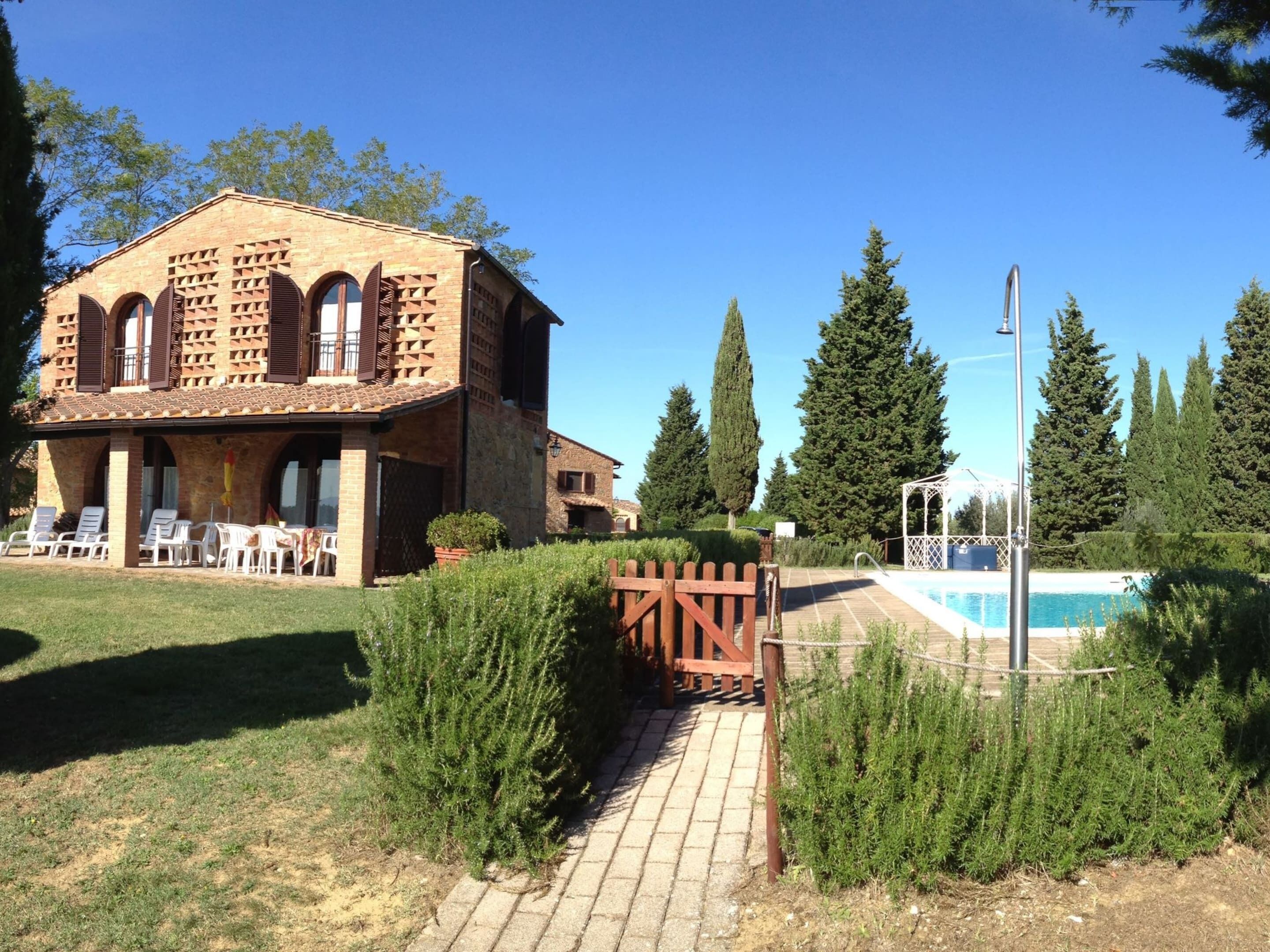 A rustic building with a brick facade and arched windows, a pool area with a wooden fence, and a clear blue sky.