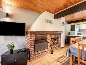 A brick fireplace with a 'HOME' sign, a TV mounted on the wall, a wooden dining table, and a kitchen area in the background. - Peaceful Farmhouse, Lessive (Rochefort)