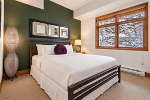 A bedroom with a bed, a nightstand, a lamp, a wall-mounted mirror, and a window with a view of trees. - Tremblant Prestige - Altitude 170-2 (Mont-Tremblant)