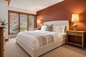 A bedroom with a large bed, two bedside tables with lamps, a wooden nightstand, and a window with blinds. - Tremblant Prestige - Altitude 170-2 (Mont-Tremblant)