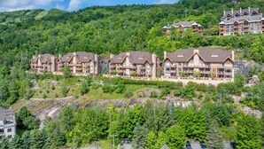 A multi-story lodge with a traditional architectural style, set against a backdrop of lush green hills. - Tremblant Prestige - Altitude 170-2 (Mont-Tremblant)