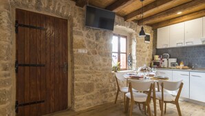A rustic stone-walled room with a wooden door, a dining table set for four, and a kitchen area with white cabinets and a flat-screen TV. - Holiday Home Near Vrana Lake and Adriatic Sea (Benkovac)