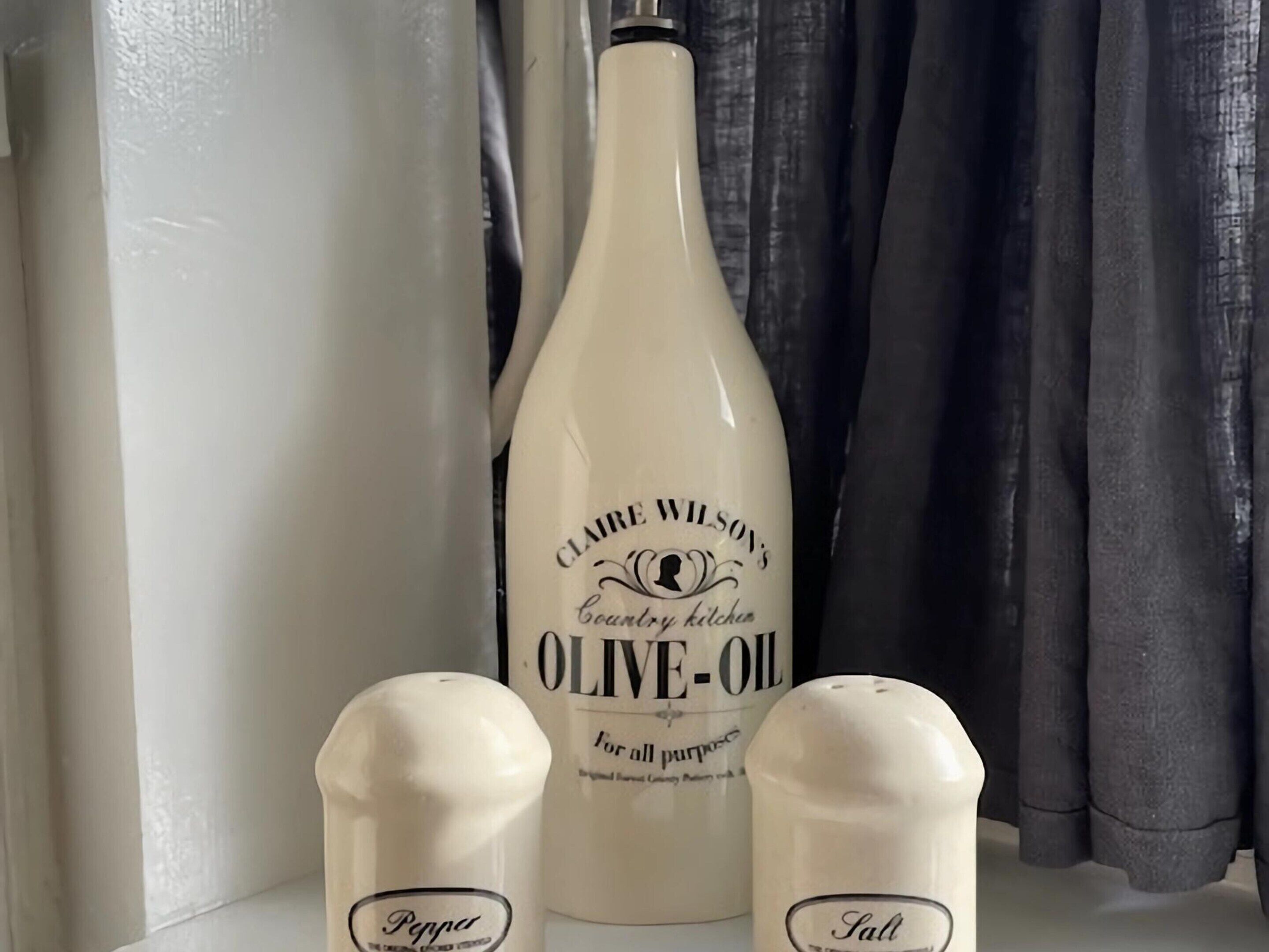 A bottle of Clare Wilson's olive oil, two salt shakers, and a pepper shaker on a shelf.