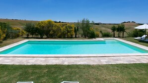 Una piscina rectangular con agua azul cristalina, rodeada por un suelo de piedra y una zona ajardinada.