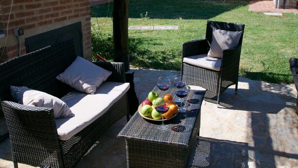 Un área de estar al aire libre con muebles de mimbre, una mesa con fruta y copas de vino, y un muro de ladrillo al fondo.