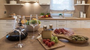 Una cocina con encimera de madera, una botella de vino, copas, un cuenco con fruta, queso y embutidos.