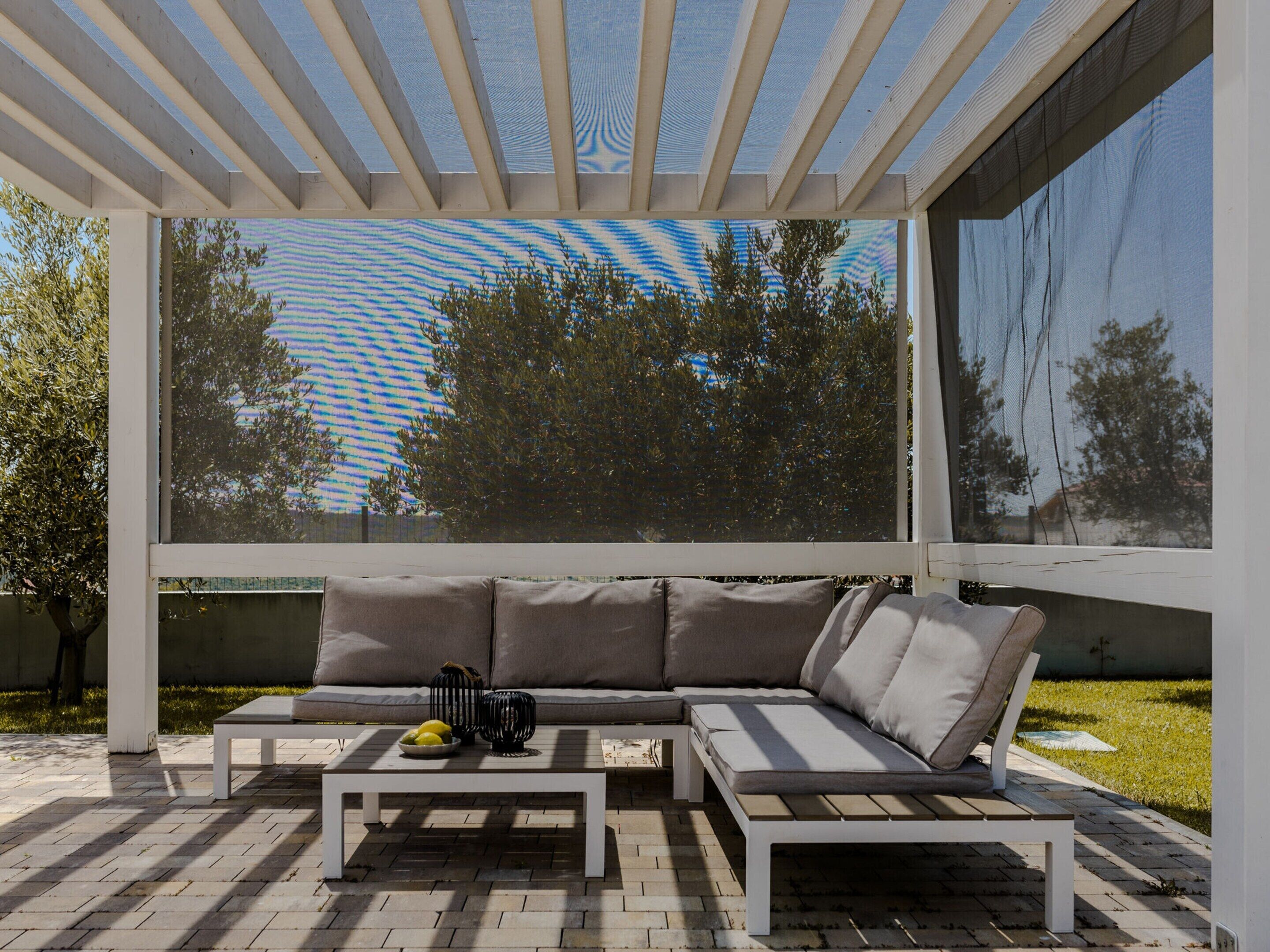 An outdoor seating area with a white bench and sofa, a coffee table, and a view of trees through a large window.