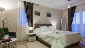 Habitación de hotel moderna con una cama grande, una mesita de noche con lámpara, una pequeña mesa auxiliar con libros, una planta en maceta y vistas al exterior a través de grandes ventanales.