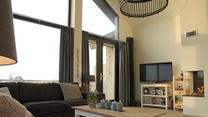 Uma sala de estar moderna com um sofá preto, uma mesa de centro de madeira e uma televisão sobre um suporte branco.