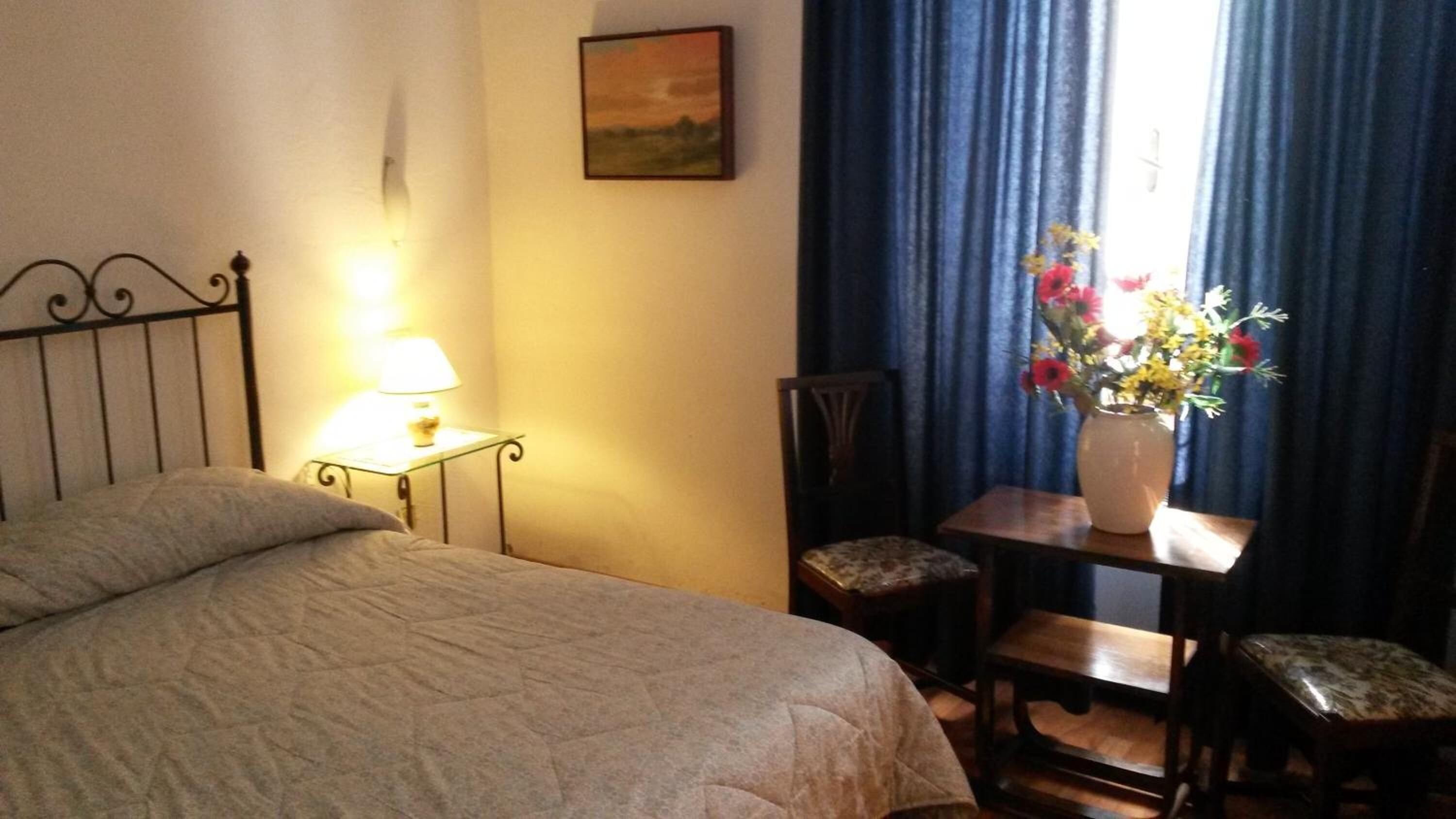Una camera da letto con un letto, un comodino con una lampada, una sedia, un tavolino con un vaso di fiori e un quadro appeso al muro.