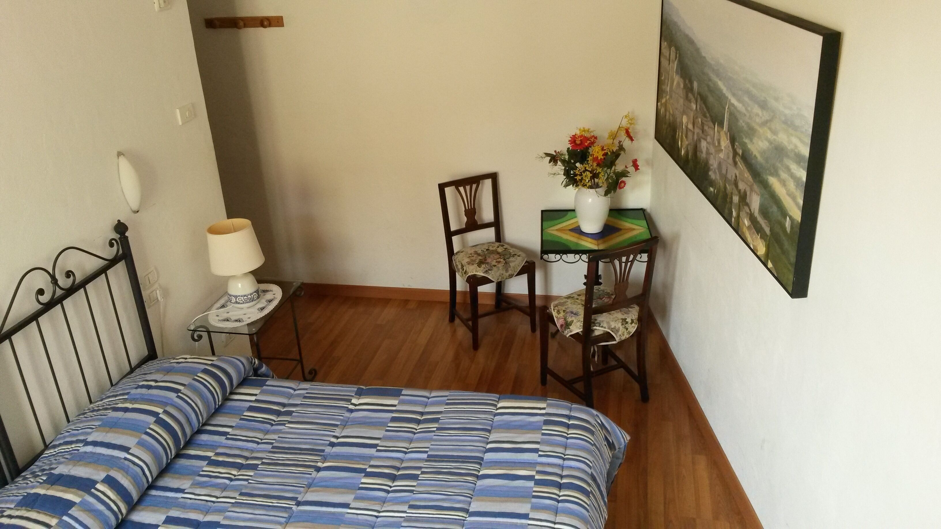 Una habitación pequeña con una cama, una silla, una mesa con un jarrón de flores y un cuadro en la pared.
