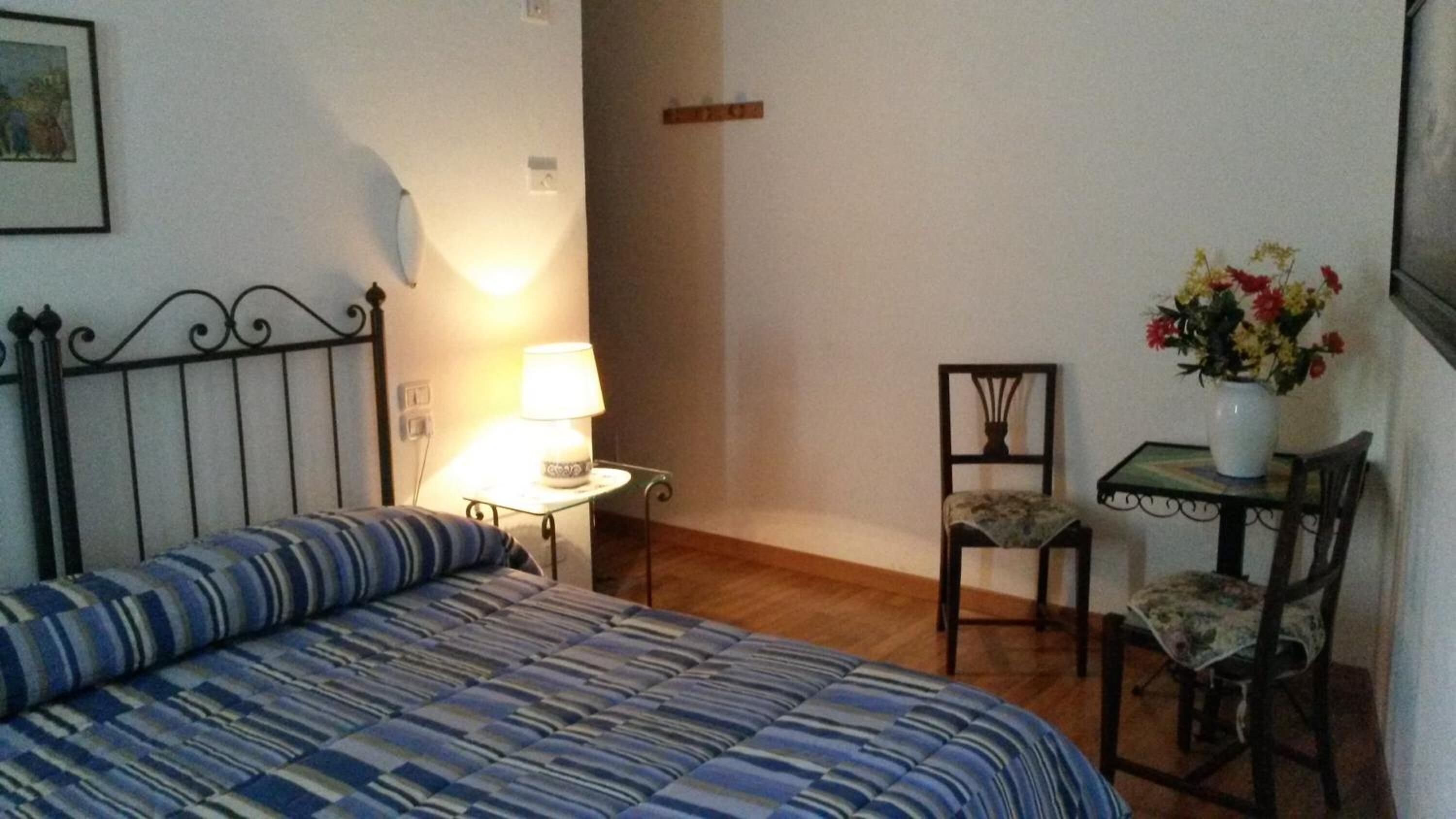 Un dormitorio con cama, una mesita de noche con lámpara, una silla y una pequeña mesa con un jarrón de flores.