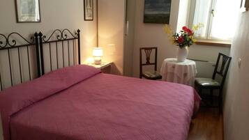 Un dormitorio con una cama grande, una mesita de noche con una lámpara, una silla, una pequeña mesa con un florero y una ventana con cortinas.
