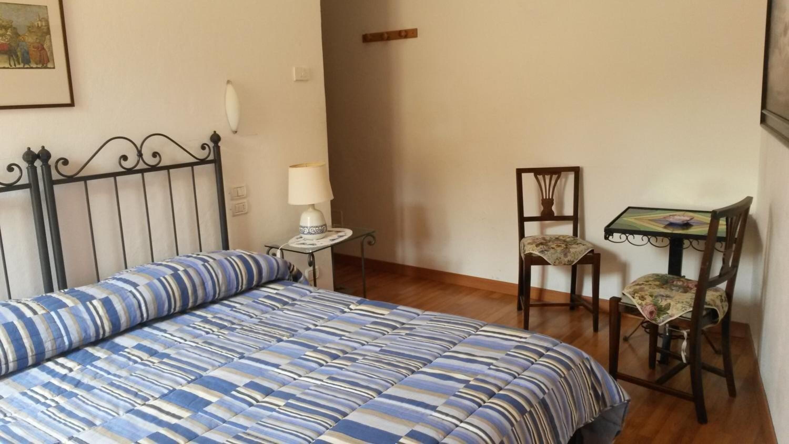A bedroom with a bed, a chair, a small table with a lamp, and a painting on the wall.