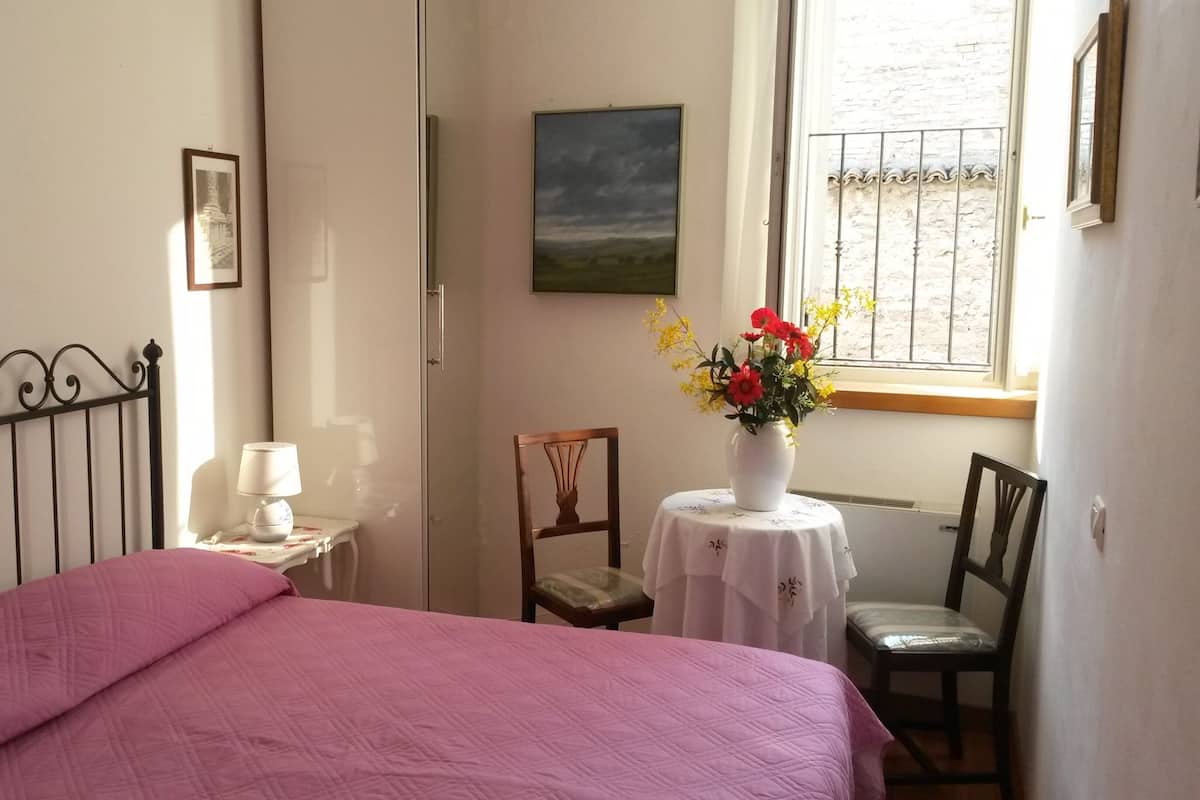 Une chambre à coucher avec un lit, une chaise, une petite table avec une lampe et un vase rempli de fleurs.