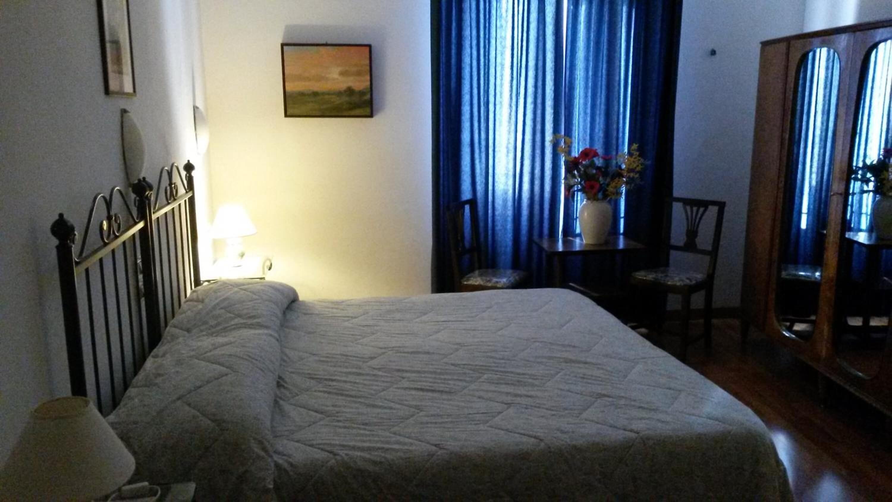 A hotel room with a bed, a chair, a table with a vase of flowers, and a painting on the wall.
