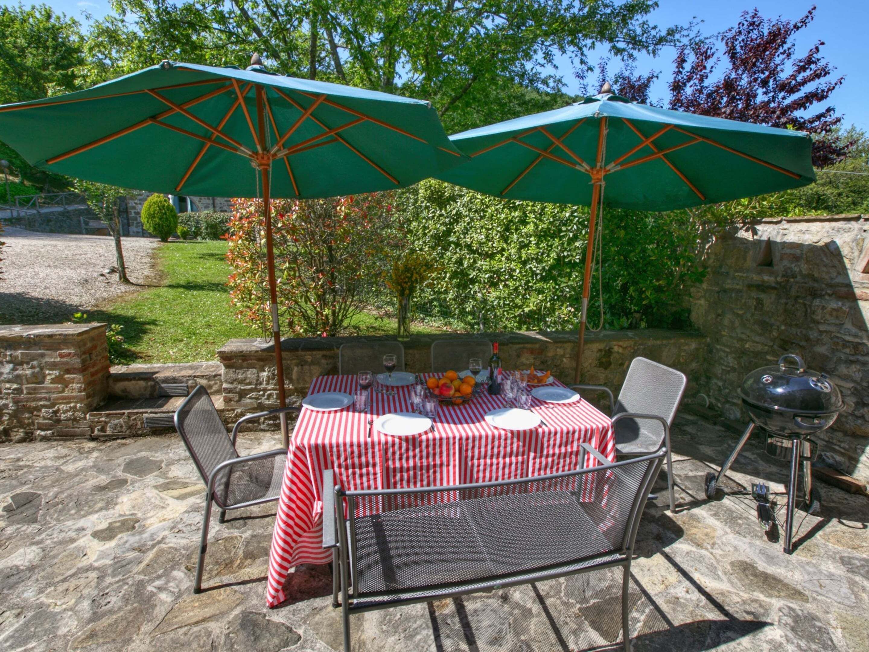 Un patio al aire libre con una mesa lista para comer, dos sombrillas verdes y una parrilla.