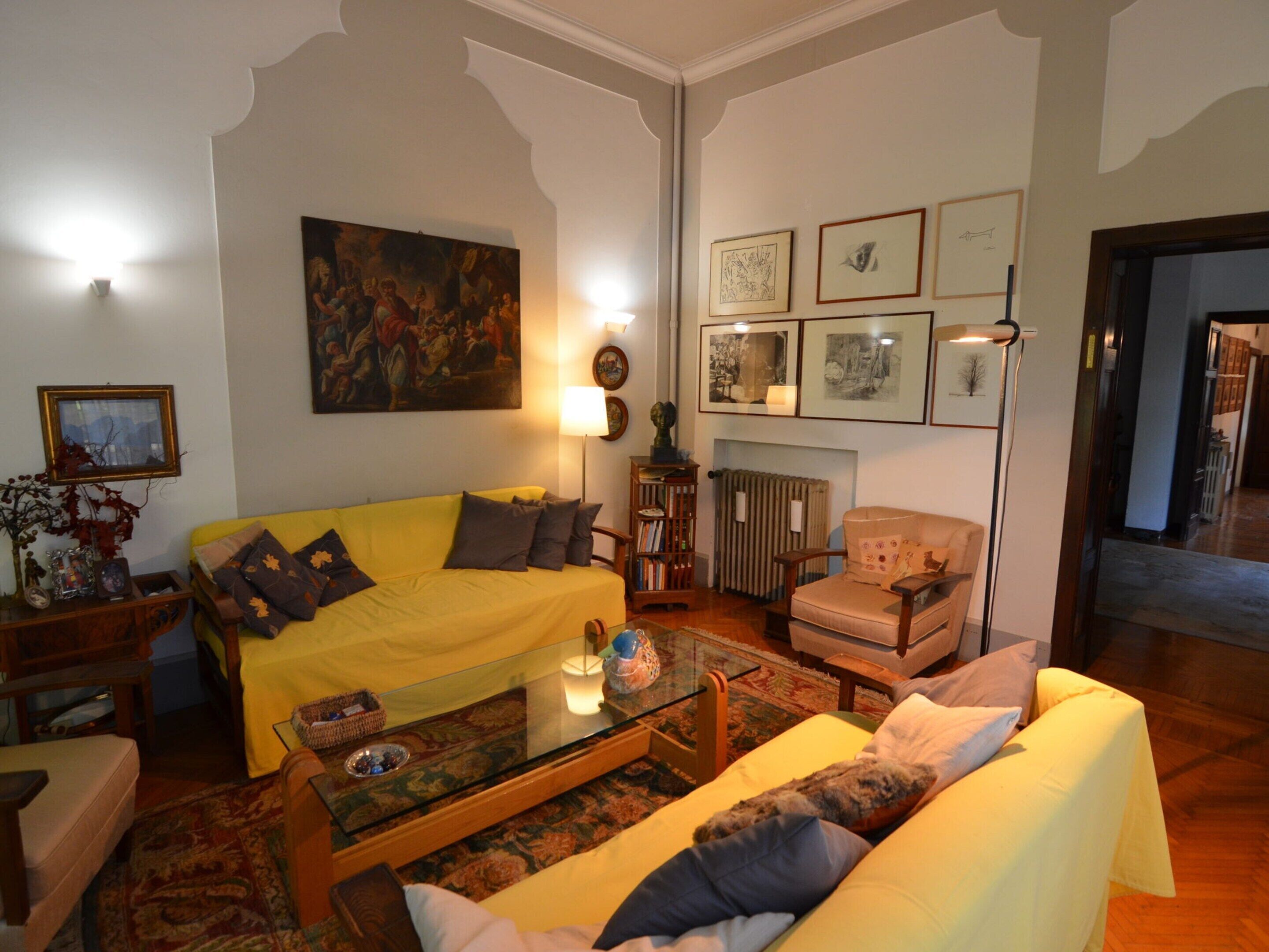 Un salón con un sofá amarillo, una mesa de centro de cristal y un sillón color crema. Hay un cuadro en la pared y varias fotografías enmarcadas.