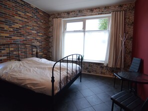 A room with a brick wall, a window with curtains, a metal bed frame, a small table, and a chair. - Home With Vinyls, Bikes and Waterways (Easterlittens)