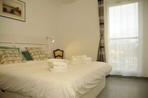 A neatly made bed with pillows and folded towels, a bedside table with a lamp, and a window with curtains.