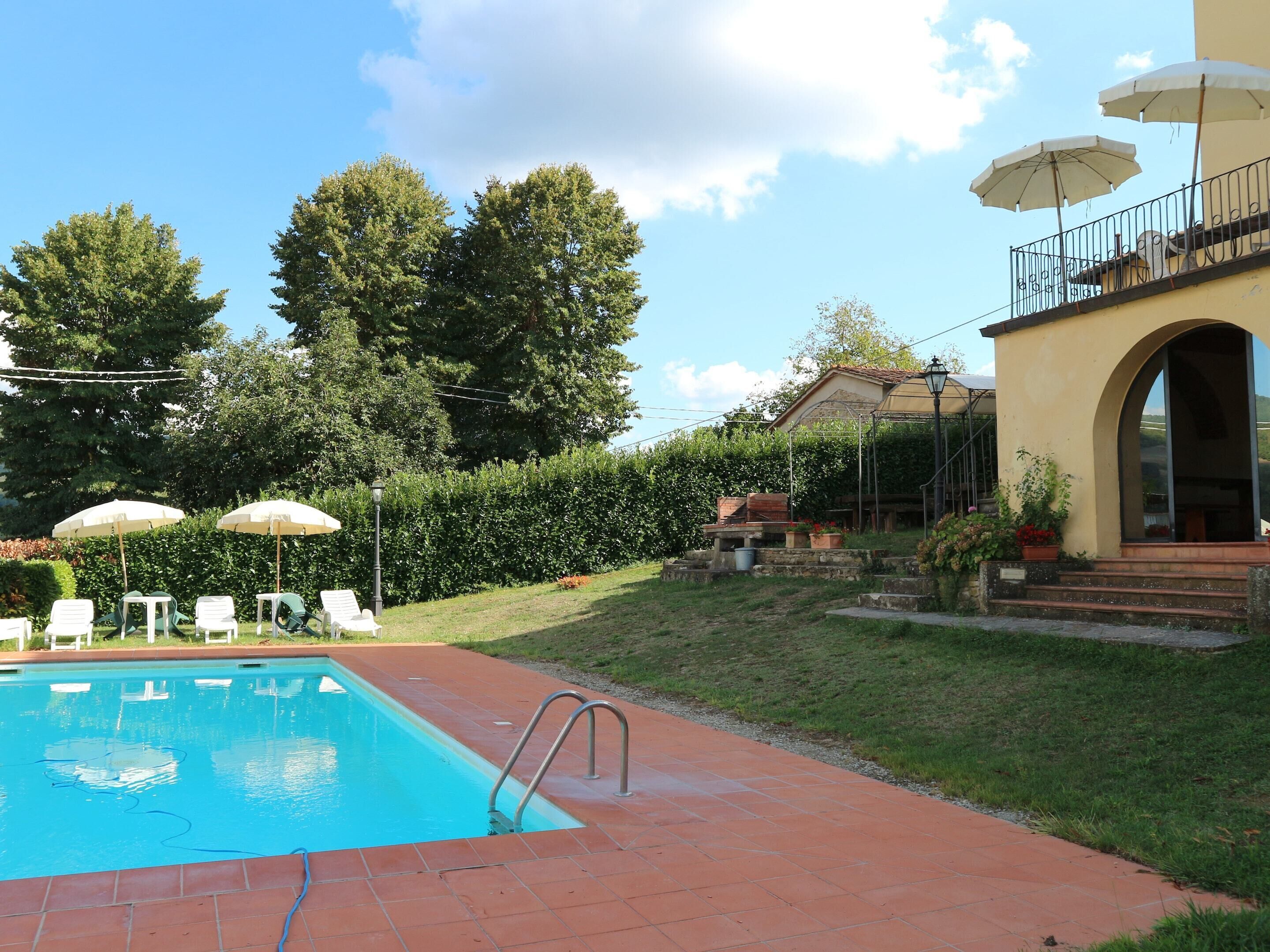 Una piscina con bordo piastrellato, circondata da sdraio e ombrelloni, con un edificio e alberi sullo sfondo.