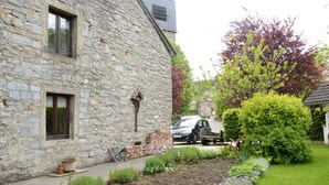 A stone building with a garden in front, a parked car, and a tree with purple leaves.