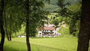 Uma casa com telhado vermelho, cercada por um campo gramado e árvores, com um pequeno lago e um caminho que a leva até ela.