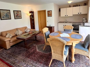 A living room with a sofa, coffee table, and wicker chairs around a round table. A kitchen with cabinets and appliances is visible in the background. - Ferienwohnung in Bruchhausen mit Balkon (Lennestadt)
