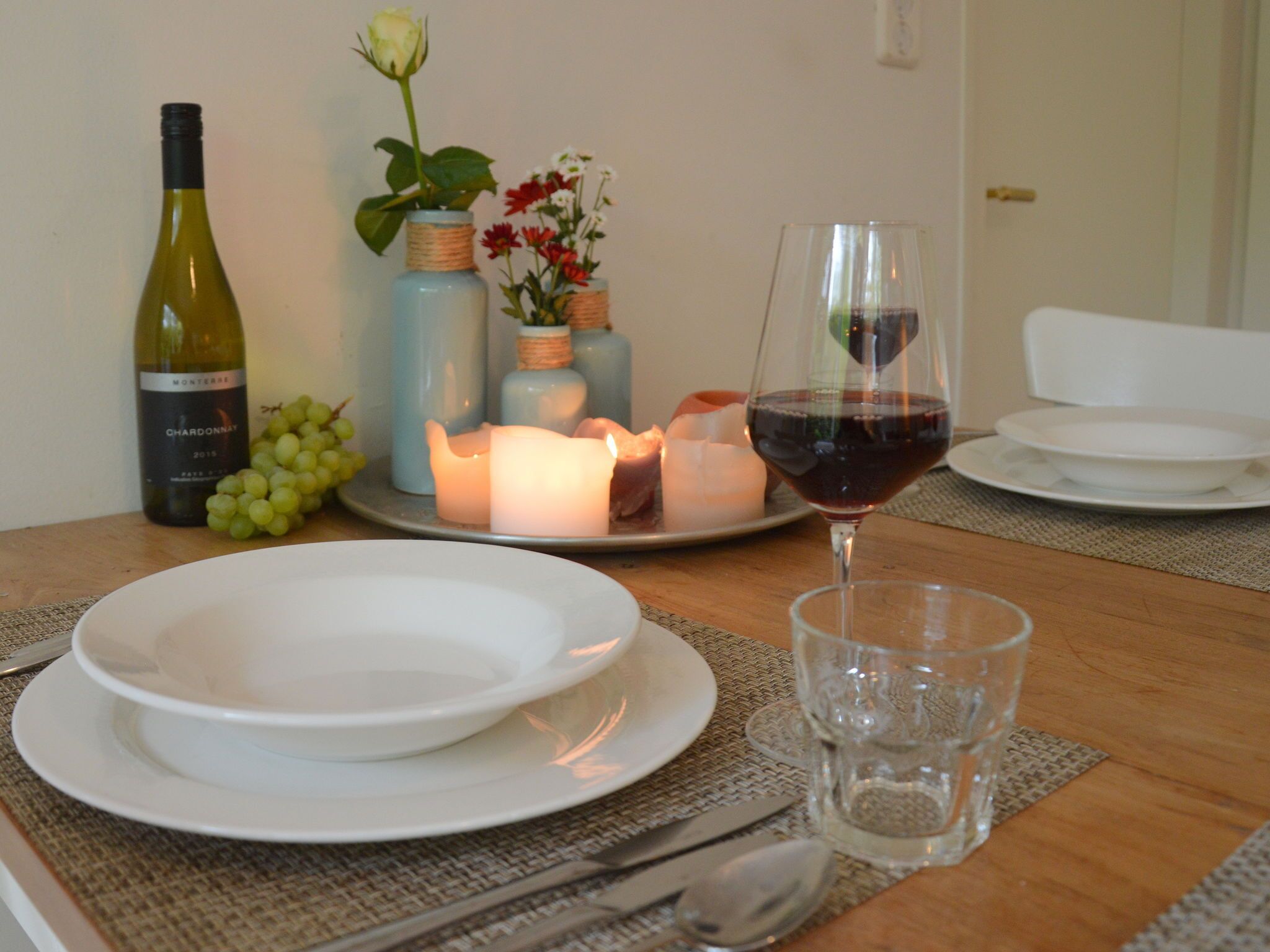 A dining table set with white plates, a glass of red wine, a bottle of chardonnay, and candles.